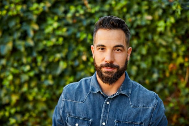 Man with dark hair and beard wearing a denim shirt standing in front of green leafy background.