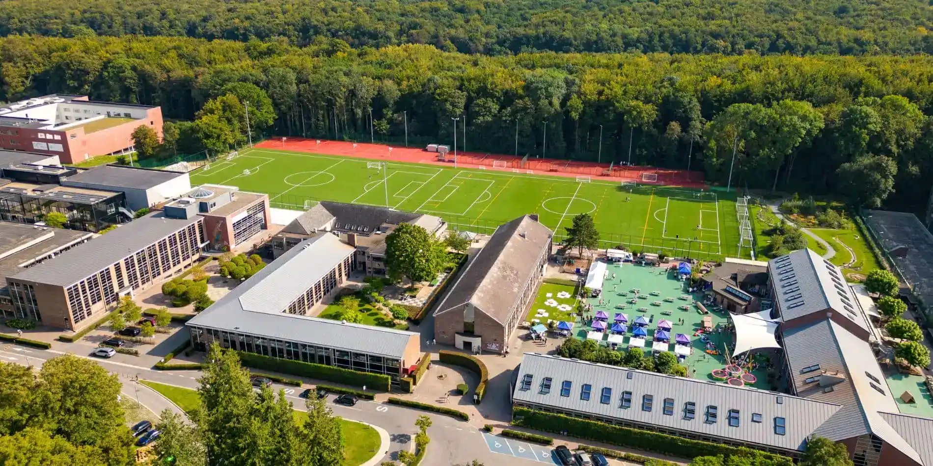 Aerial view of a school campus with multiple buildings, a large soccer field, and a courtyard featuring tents and outdoor seating, bordered by a dense forest.