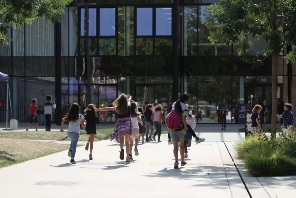 Group of children running and walking on a paved path outside a modern building with large glass windows.