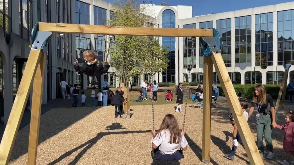 Children playing and swinging on wooden swings in a school playground with modern glass buildings in the background.