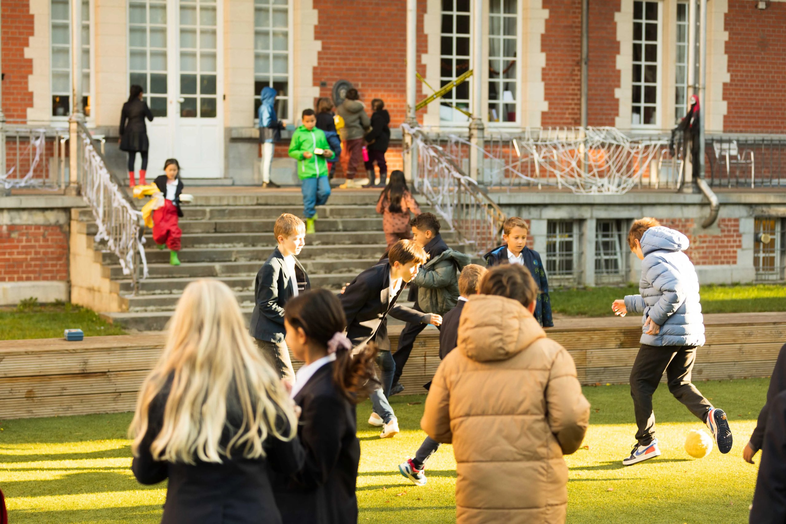 Children playing soccer on a grassy field in front of a brick building with stairs during daytime.
