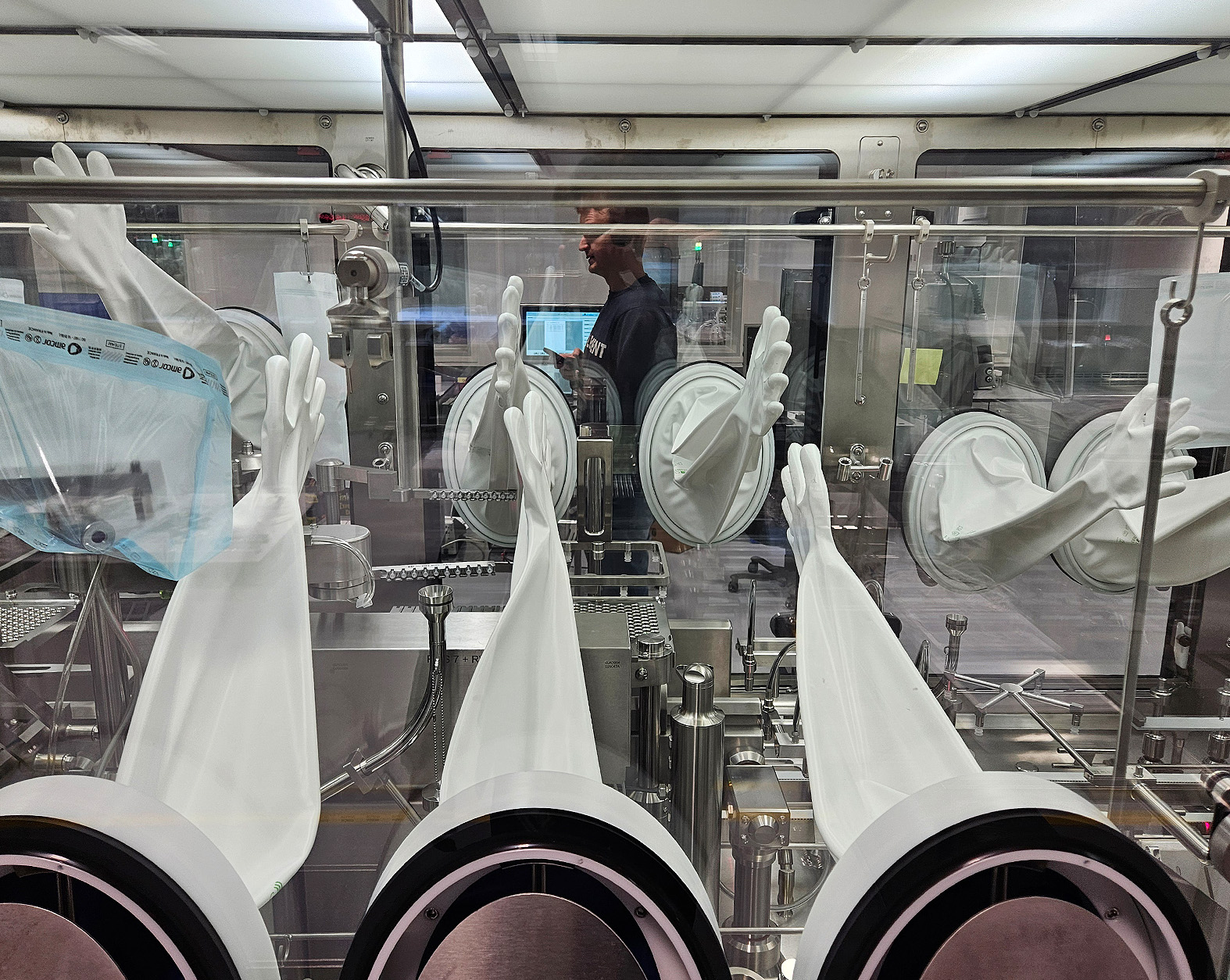 Aseptic isolator with white gloves attached, used for sterile pharmaceutical filling, with a reflection of a person working on a computer behind the glass.