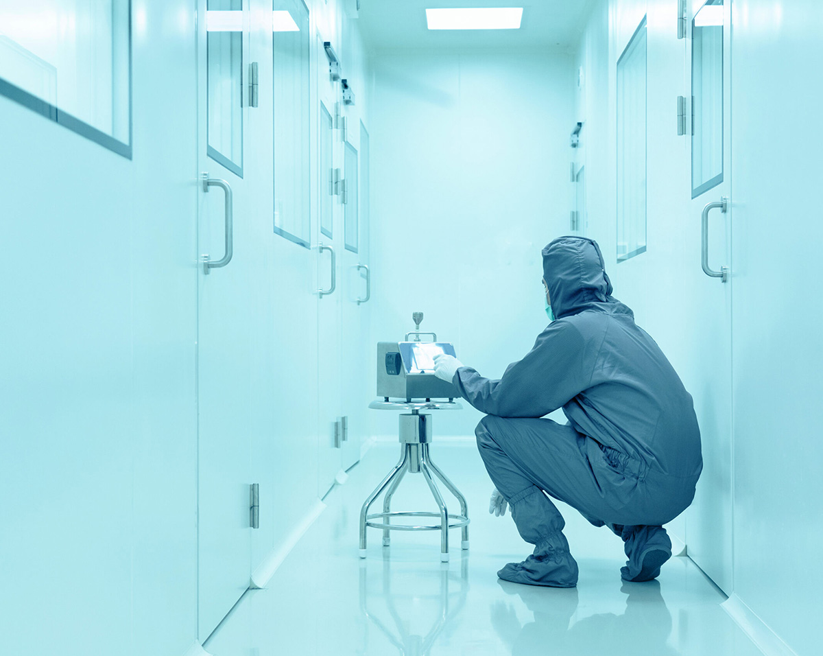 Person in full protective suit squatting in a cleanroom hallway interacting with a device on a stool.