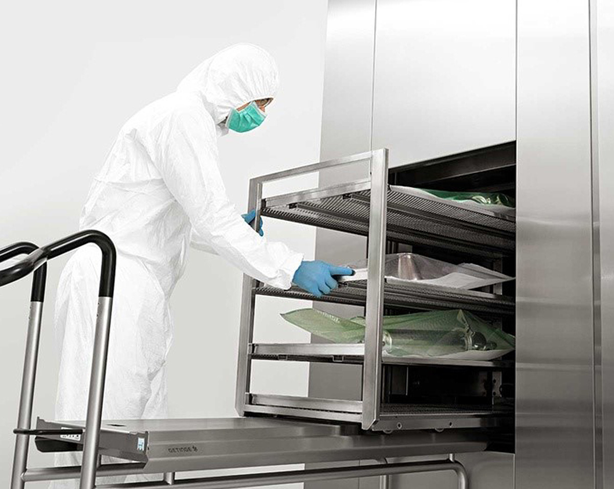 Person in white protective suit, face mask, and gloves handling a stainless steel rack in a sterilization machine.