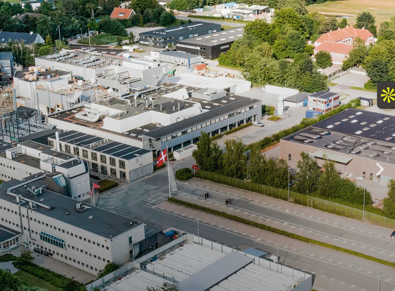 Aerial view of a large industrial or research facility with multiple buildings, parking lots, and green trees surrounding the area, featuring a Danish flag on a flagpole.