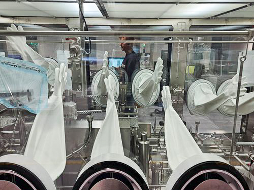 Aseptic isolator with white gloves attached, used for sterile pharmaceutical filling, with a reflection of a person working on a computer behind the glass.
