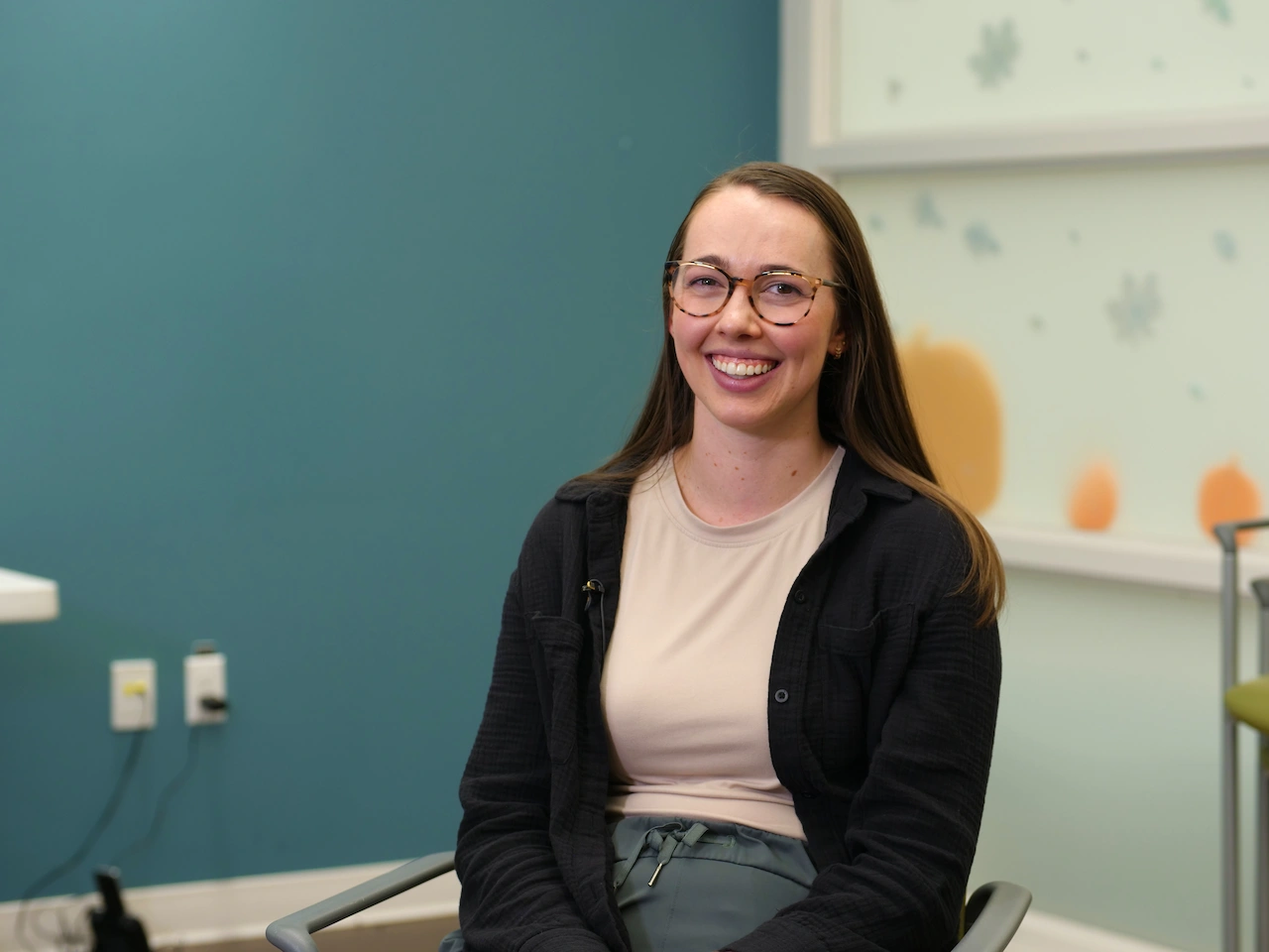 Smiling woman with glasses seated in a chair against a teal and light wall with decorations.