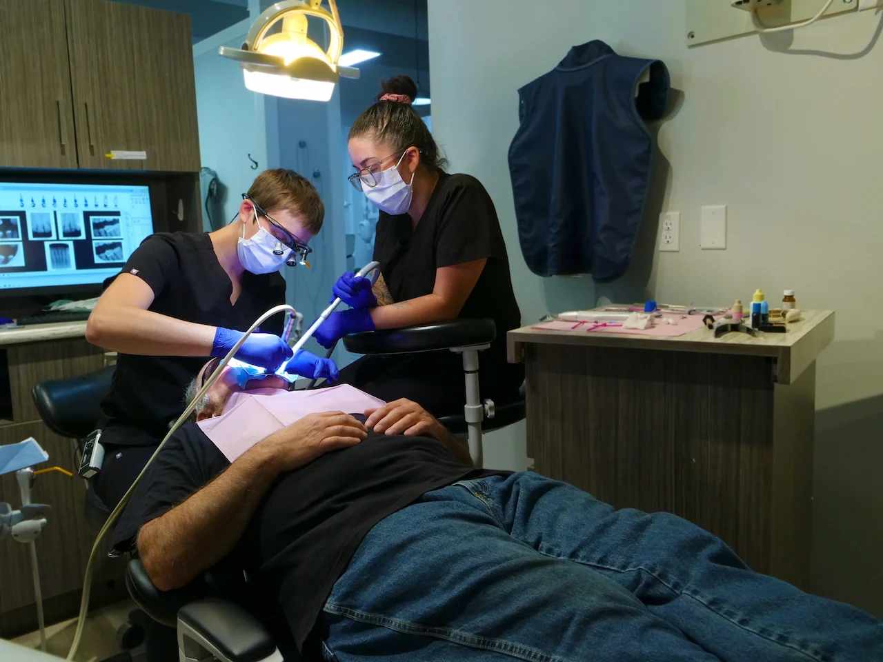 Two masked dental professionals treating a patient lying on a dental chair under a light.