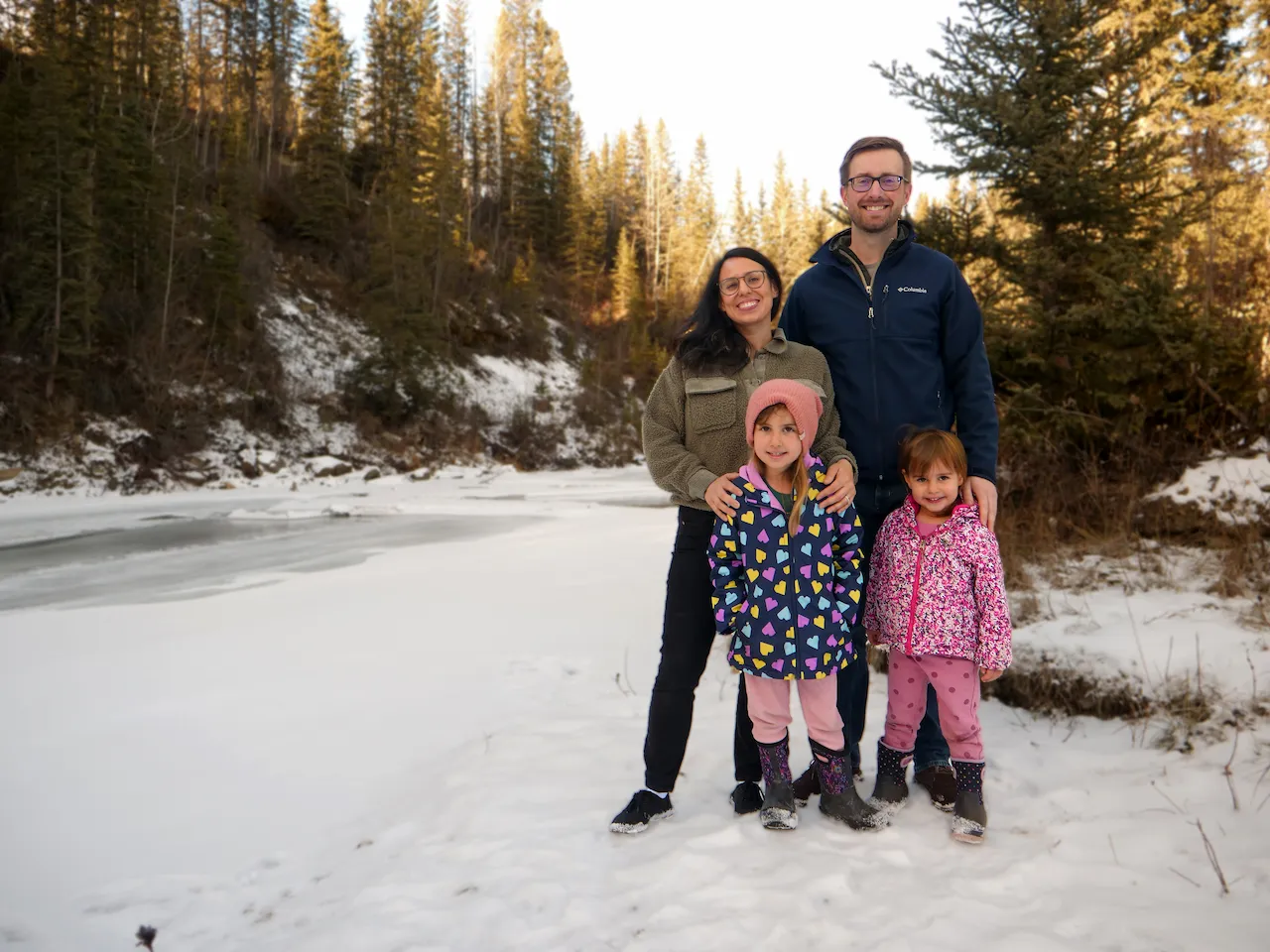 Smiling family of four standing on snowy ground near a frozen river with evergreen trees in the background.