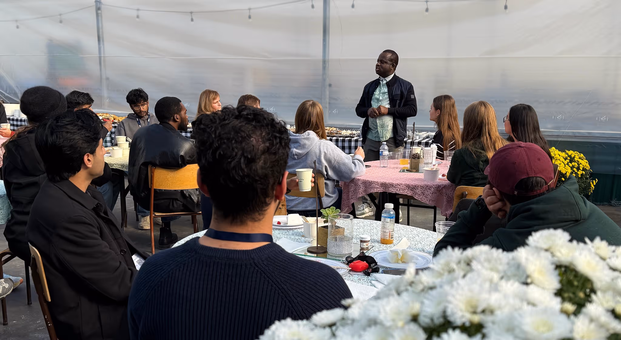 Diverse group seated at tables listening to a standing speaker in a covered outdoor setting.