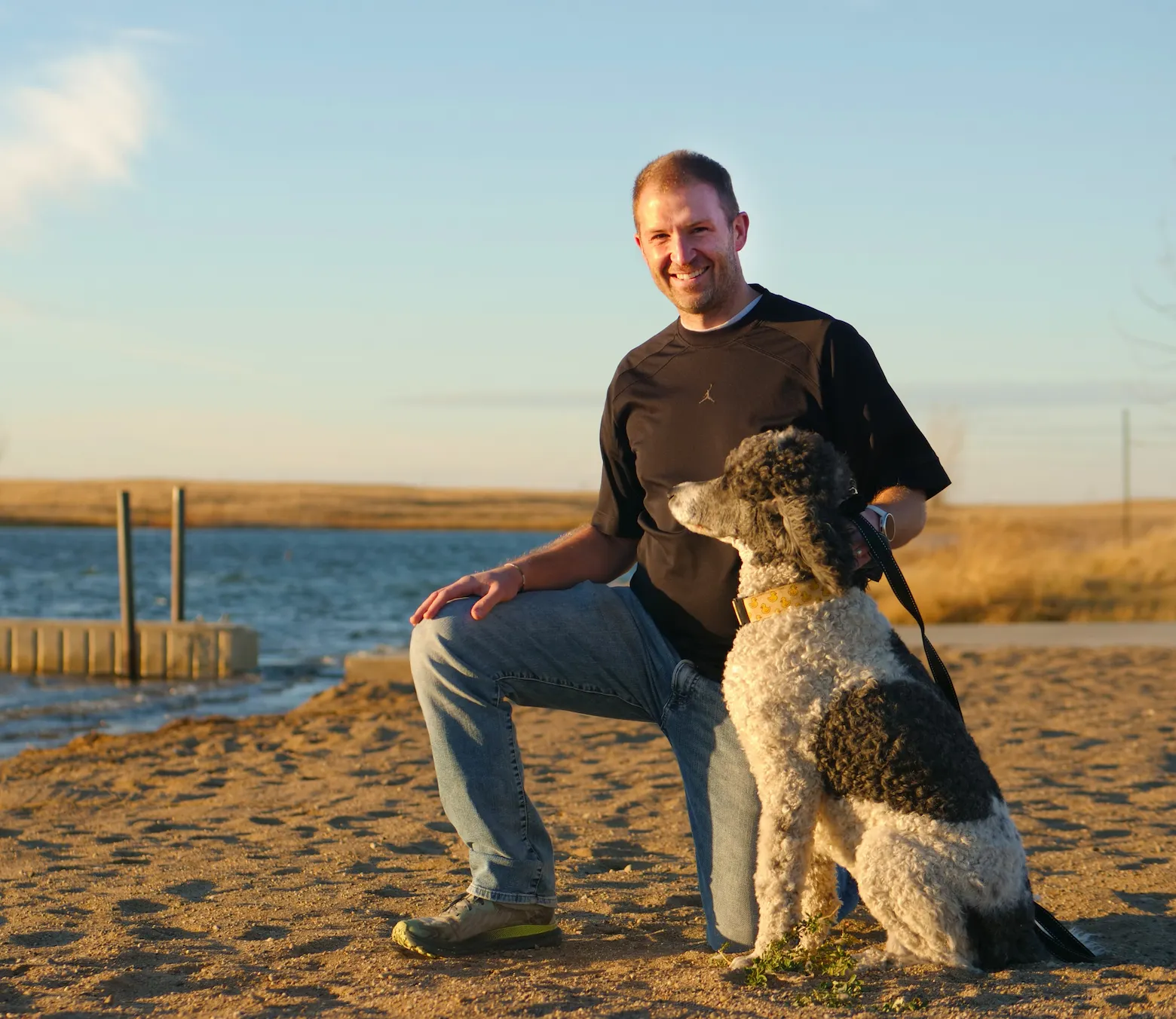Man in black shirt kneels on sandy shore with a black and white curly dog beside him near a lake.