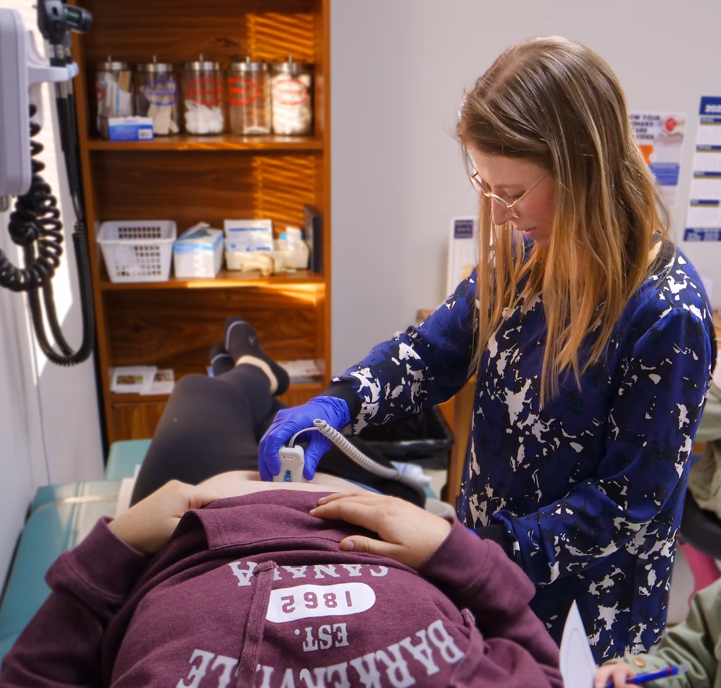 Medical professional wearing glasses and blue gloves using an ultrasound device on a patient's abdomen in a clinic.