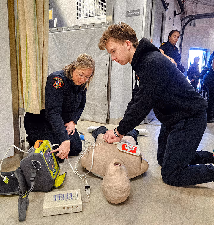 Two people practicing CPR and defibrillator use on a training mannequin indoors.