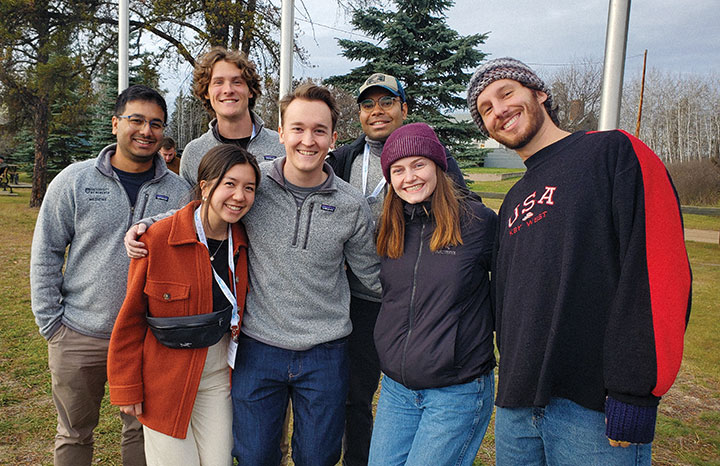 Group of seven young adults smiling outdoors with trees and grass in the background.