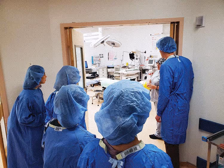 Medical team in blue scrubs and caps preparing outside an operating room with surgical equipment inside.