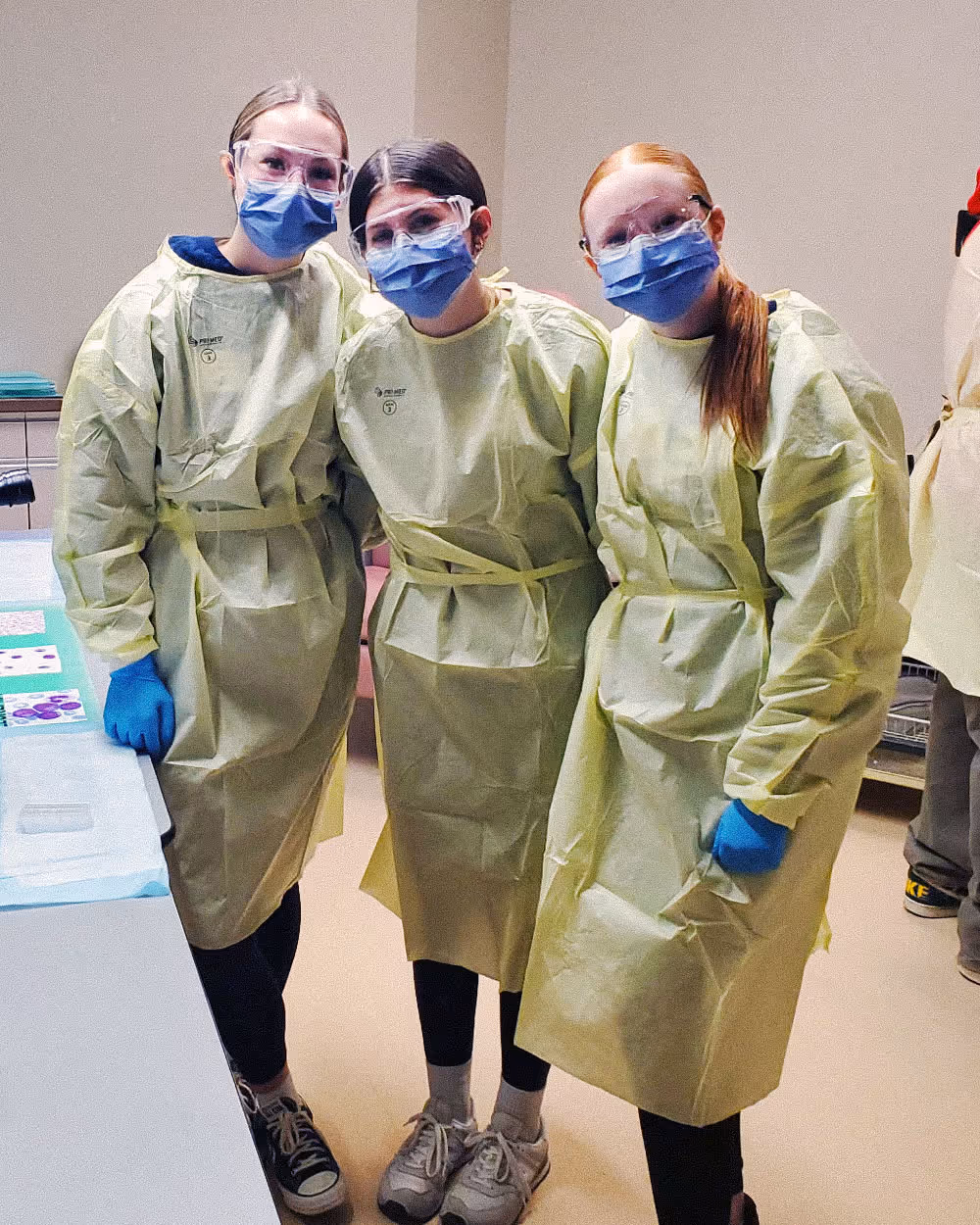 Three people wearing yellow protective gowns, blue face masks, gloves, and clear goggles indoors.