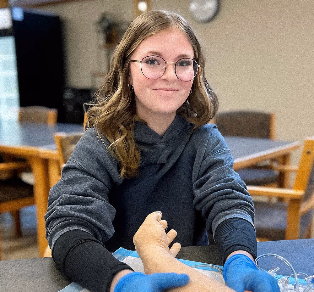 Smiling young woman with glasses and gloves drawing blood from a patient's hand in a clinical setting.