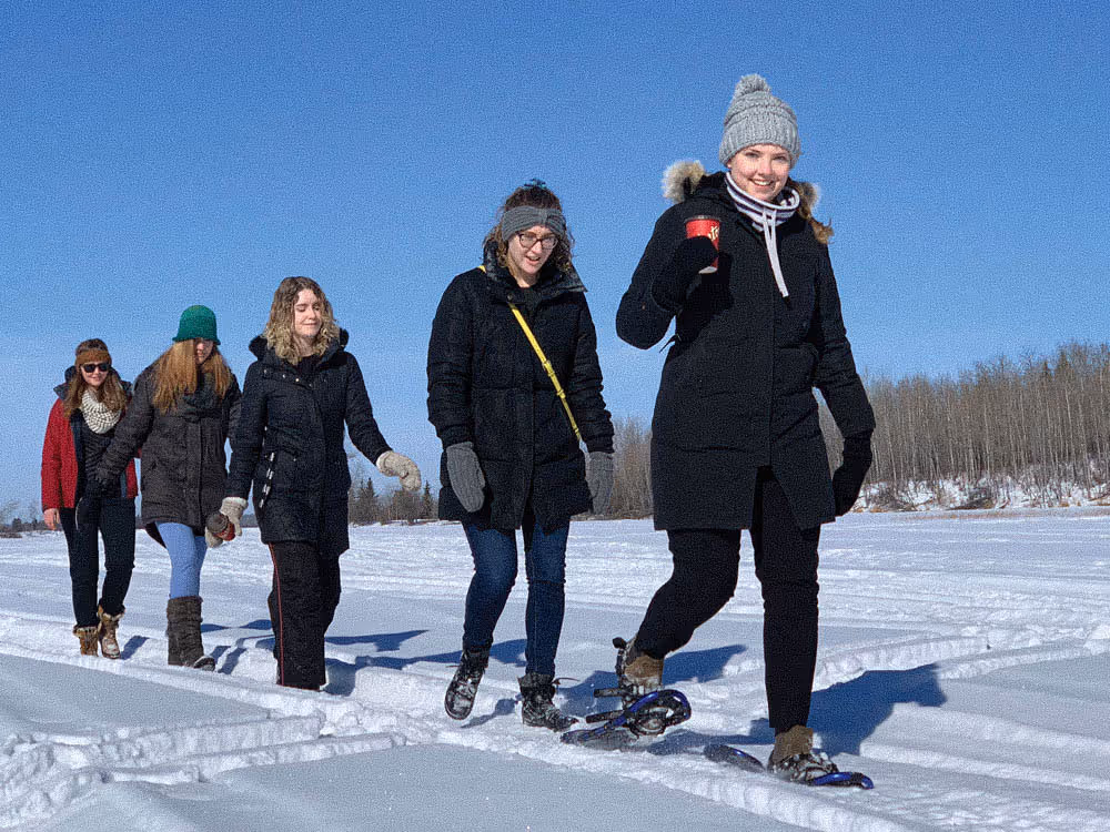 Group of five women walking in snow wearing winter clothes and snowshoes under clear blue sky.