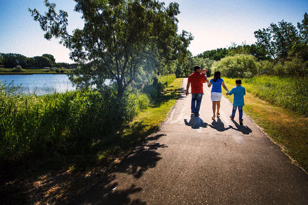 Family of four walking on a sunny path near a lake with trees and grass.