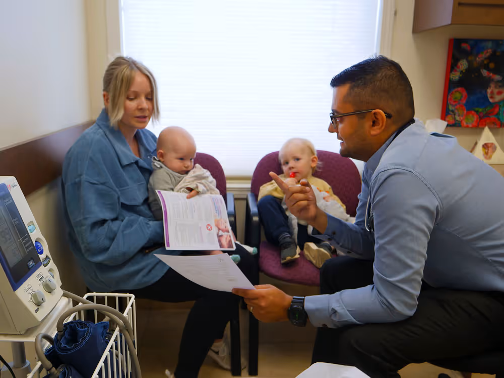 Doctor talks with mother holding infant and toddler sitting nearby in a medical office.