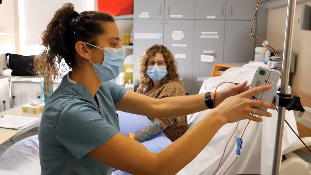 Nurse in mask adjusting IV pump while patient with tattoo and mask watches in hospital room.
