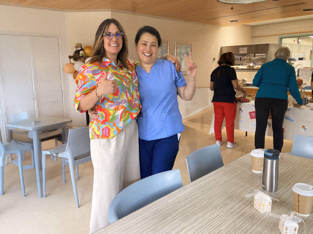Two women smiling, one in colorful shirt giving thumbs up, the other in scrubs showing peace sign.