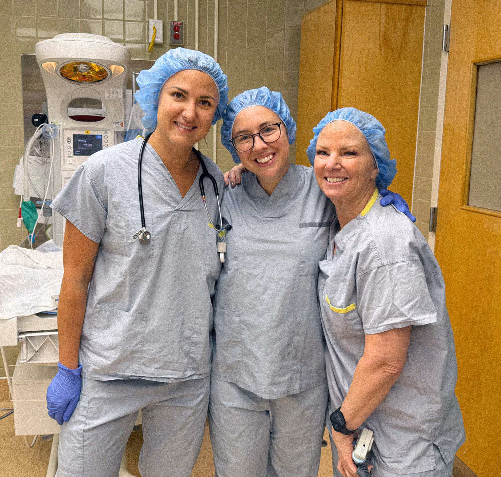 Three medical professionals in scrubs and blue caps smiling in a hospital room.