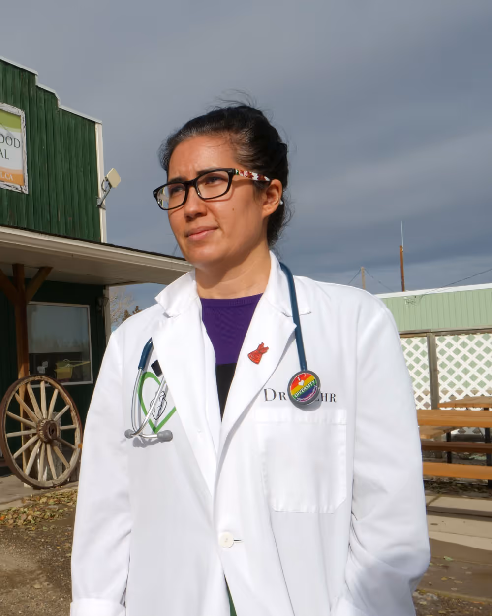 Female doctor wearing glasses and white coat with stethoscope outdoors near a green building.