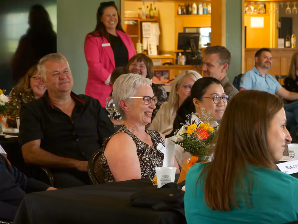 Group of people smiling and listening at an indoor event with flowers on the table.