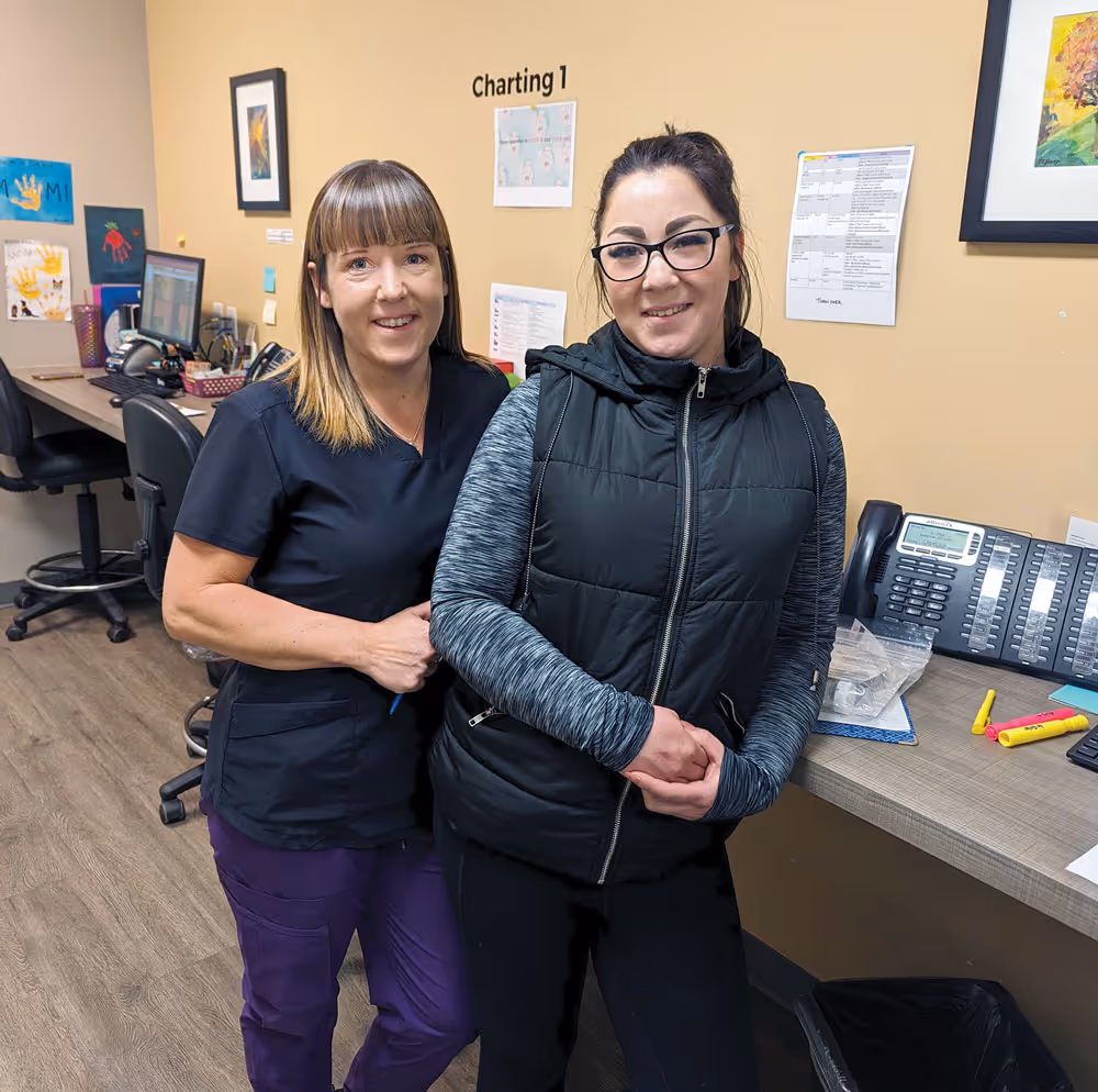 Two women smiling in an office; one wears scrubs, the other a black vest and glasses.