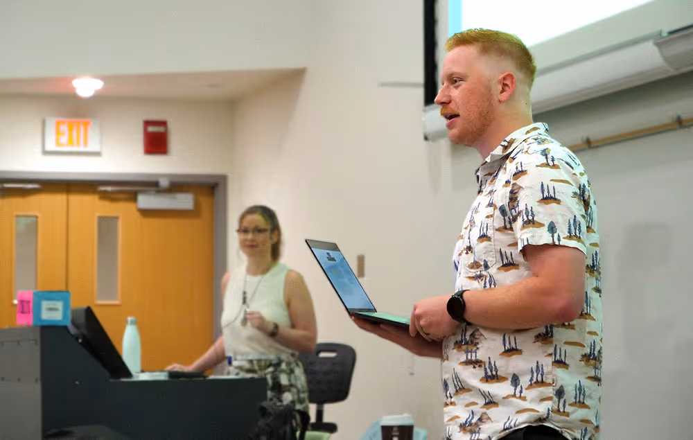 Man holding laptop speaking in classroom with woman standing near podium in background.