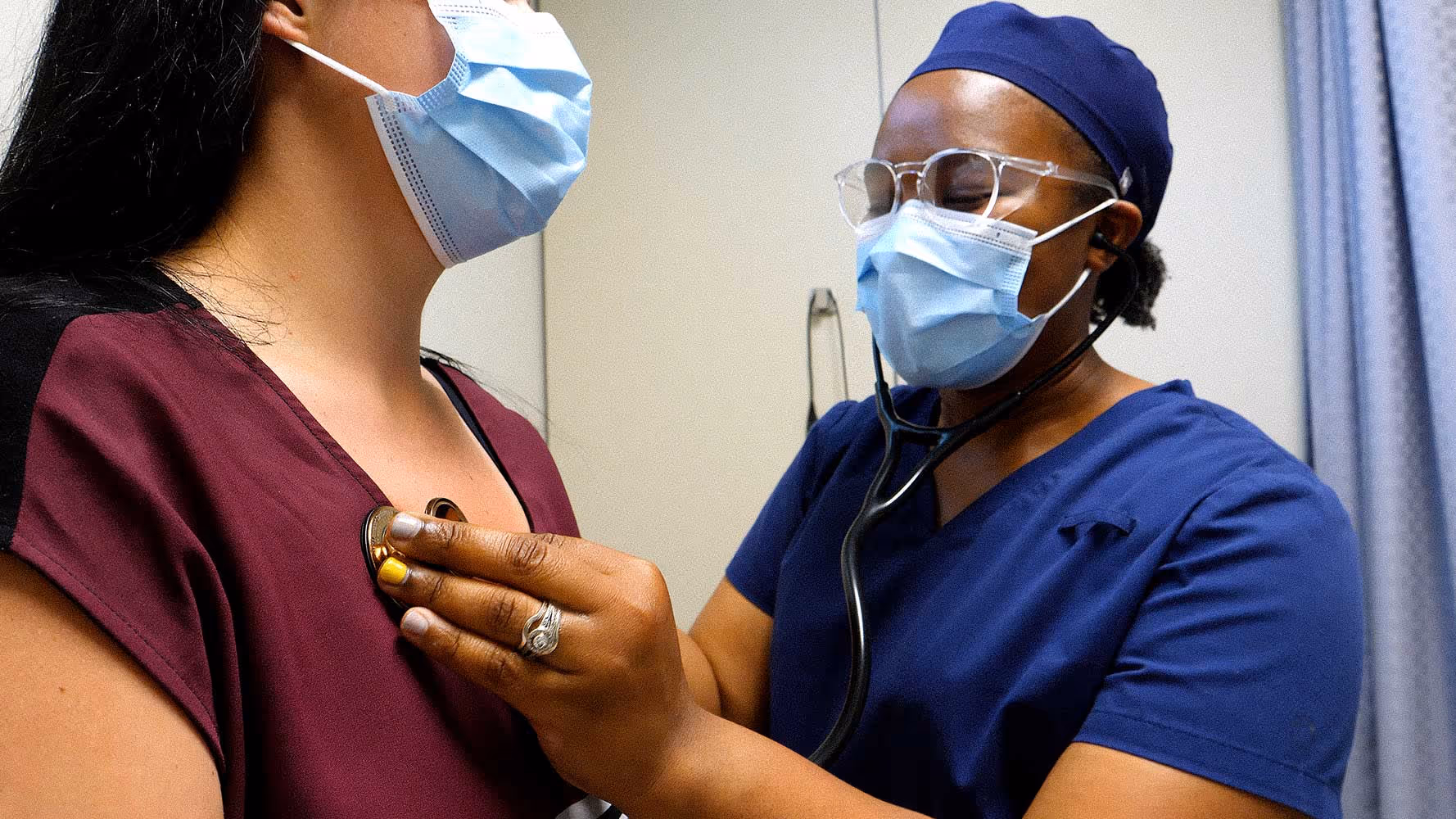 Healthcare worker in blue scrubs uses a stethoscope to listen to a masked patient's chest.