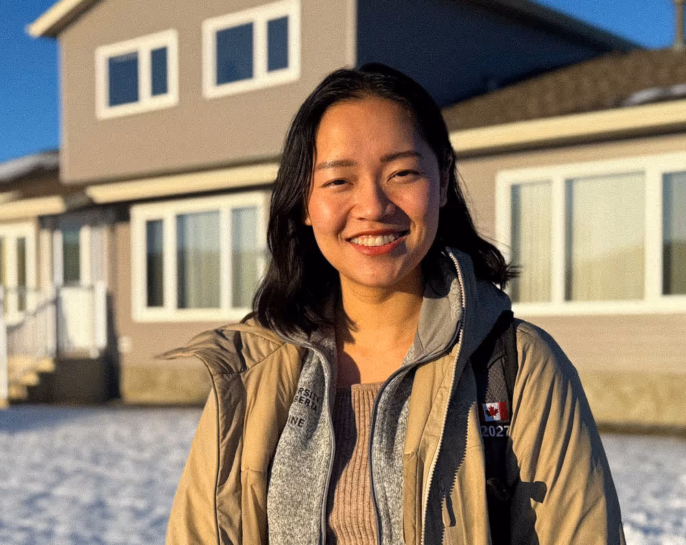 Smiling woman in a beige jacket standing outside in sunlight with a building and snow in the background.