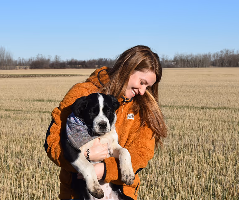 Young woman in an orange jacket smiles while holding a black and white dog in a field.