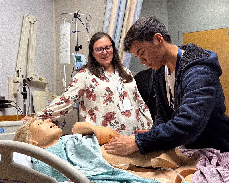Young man practicing medical exam on a hospital mannequin while woman watches in clinical room.