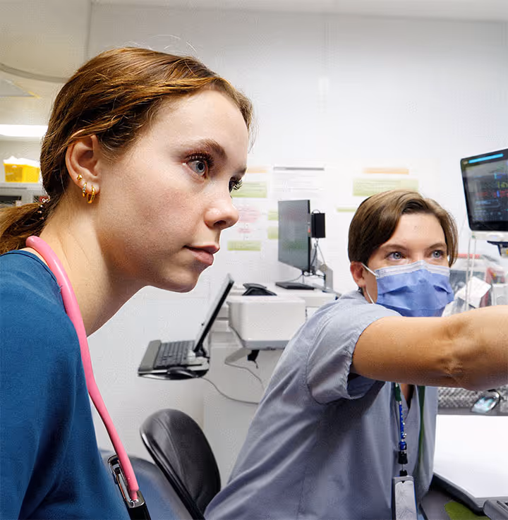 Two healthcare workers in a clinical setting, one wearing a face mask pointing at a monitor, the other with a stethoscope.