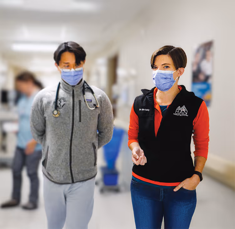 Two masked healthcare professionals, a man with a stethoscope and a woman in a black vest, walking in a hospital.