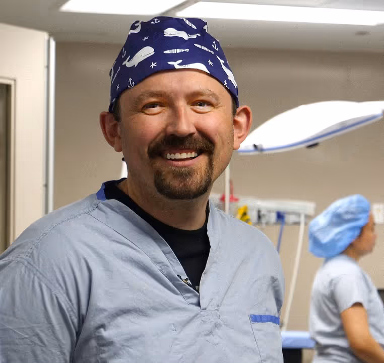 Smiling male medical professional in surgical scrubs and whale-patterned cap in a hospital room.
