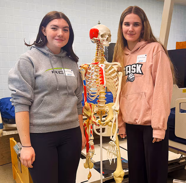 Two girls standing indoors on each side of an anatomical skeleton model with muscle highlights.