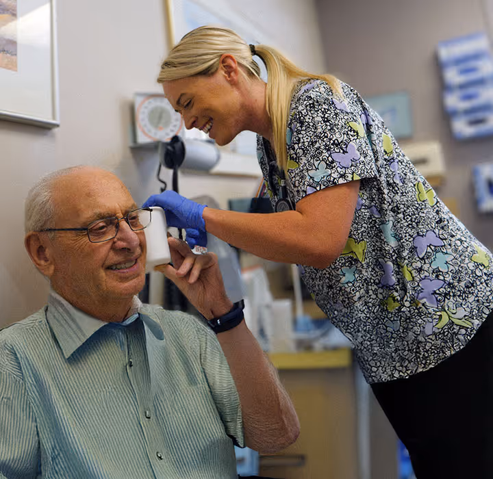 Healthcare worker wearing gloves assists elderly man by placing hearing aid in his ear.