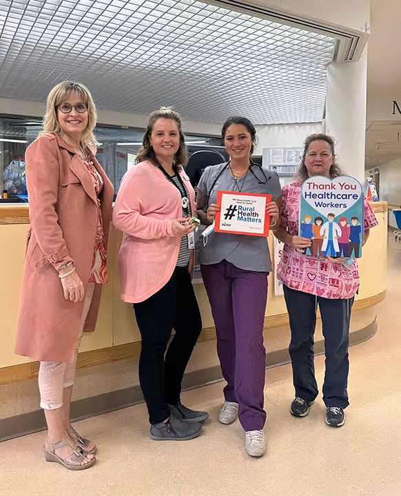 Four smiling healthcare workers indoors, two holding signs: 'Rural Health Matters' and 'Thank You Healthcare Workers'.