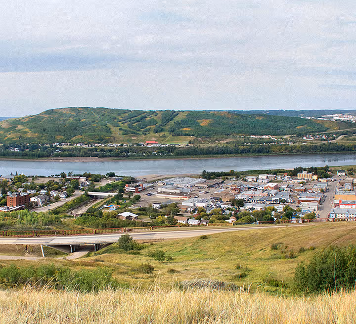 Town with houses, roads, and a river with green hills in the background under a cloudy sky.