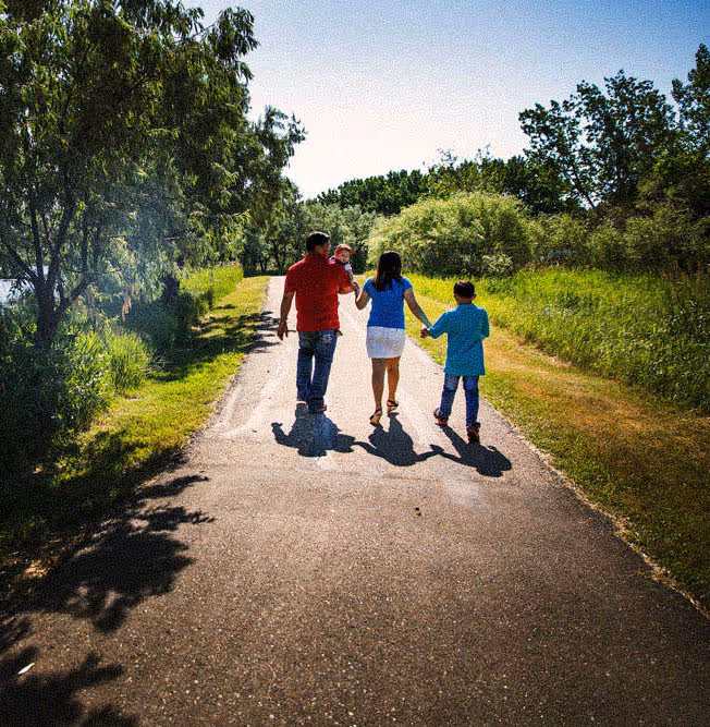 Family of four walking on a sunny paved path surrounded by green trees and grass.