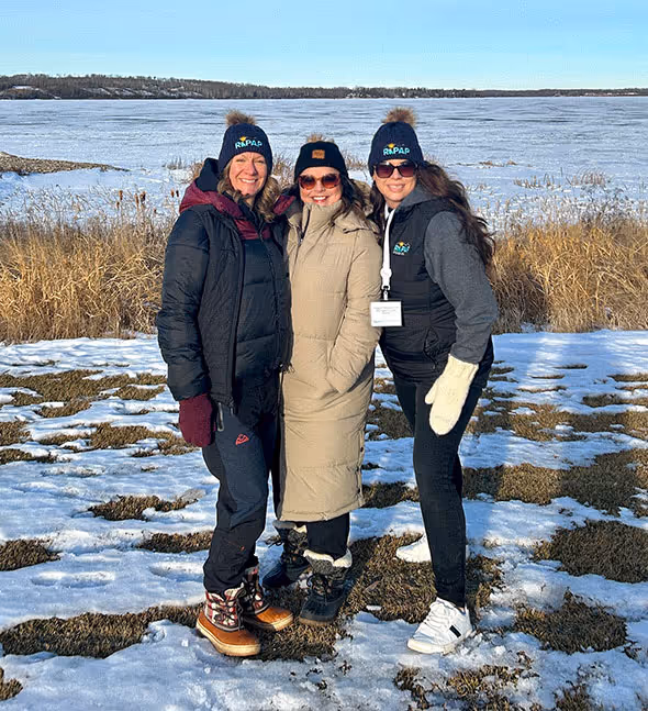 Three women wearing winter coats, hats, and boots posing outside on snow-covered ground near a frozen lake.