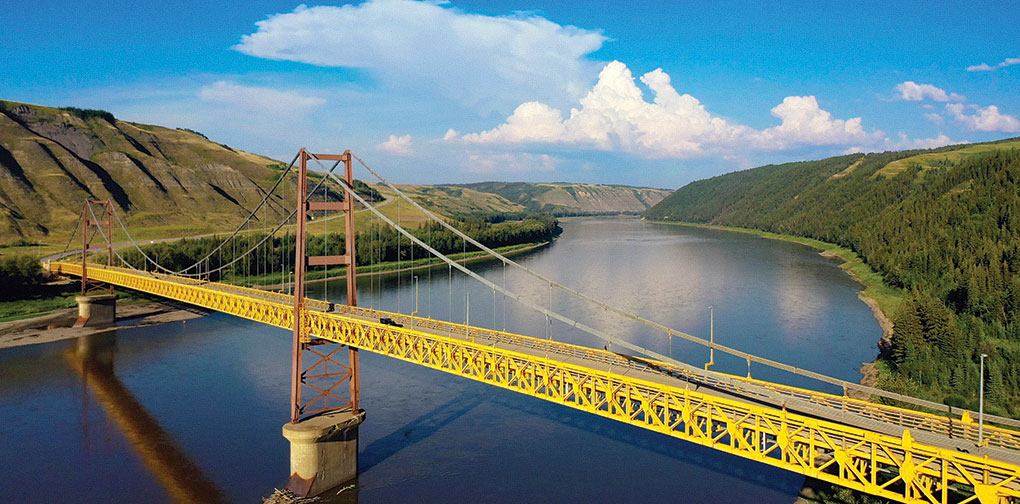Yellow suspension bridge spanning a wide river with green hills and blue sky with clouds.