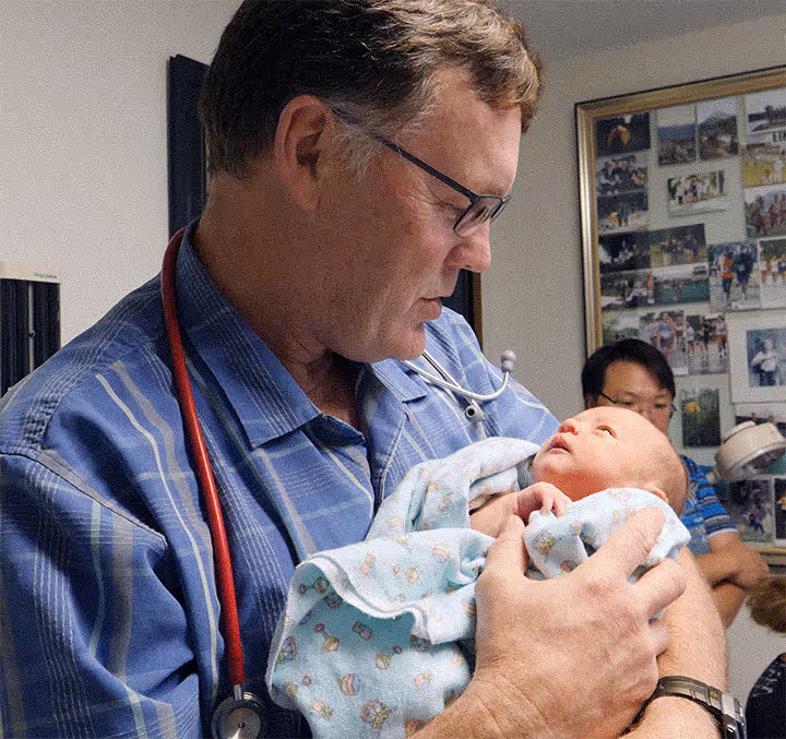 Male doctor with stethoscope holding and examining a newborn baby wrapped in a blue blanket.