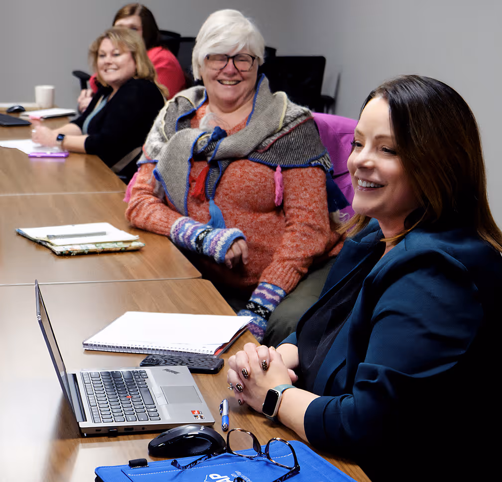 Four women sit at a conference table with laptops, notebooks, and coffee, smiling and engaged in a meeting.