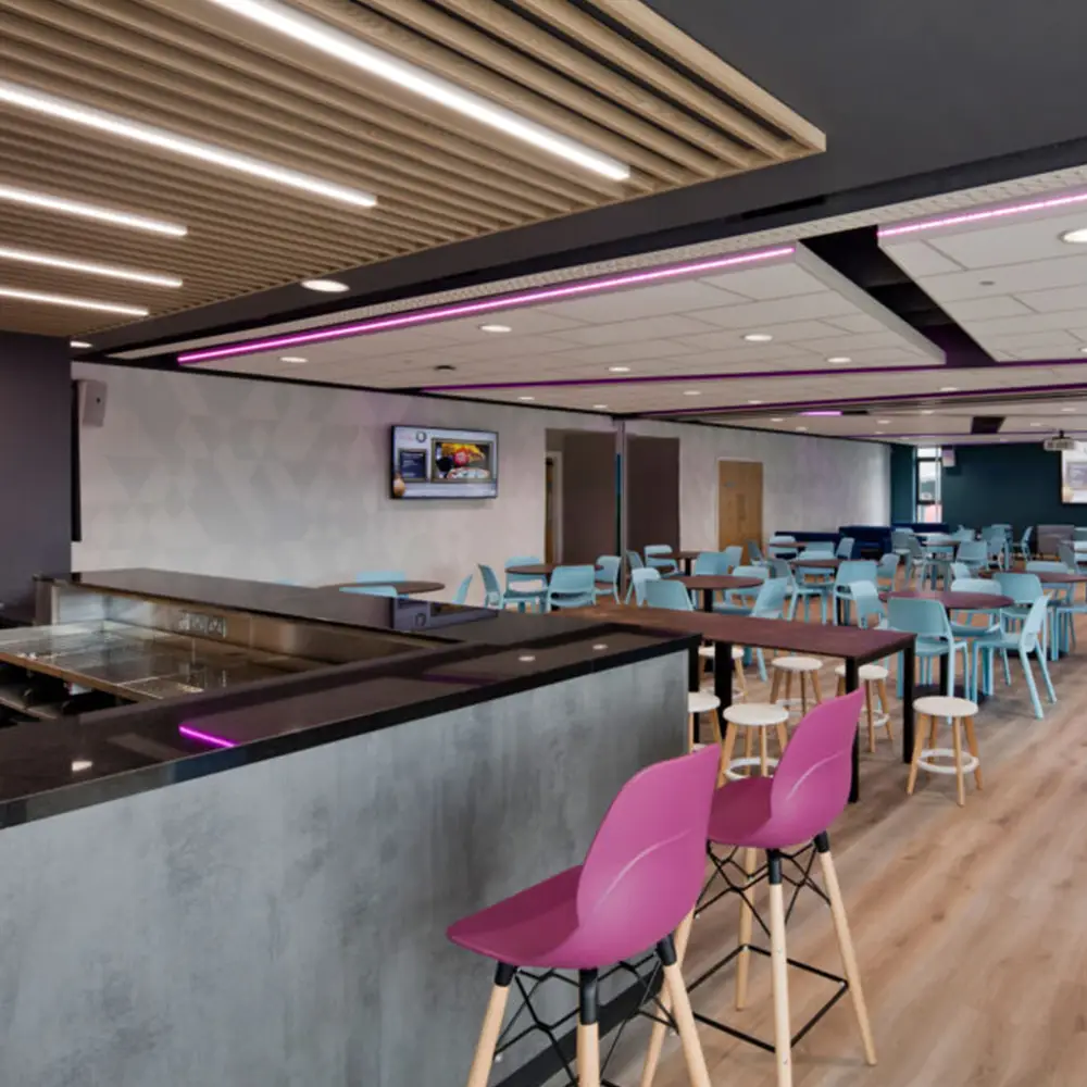 Modern cafeteria with purple bar stools, blue chairs, wooden tables, and wood floor with purple ceiling lights.
