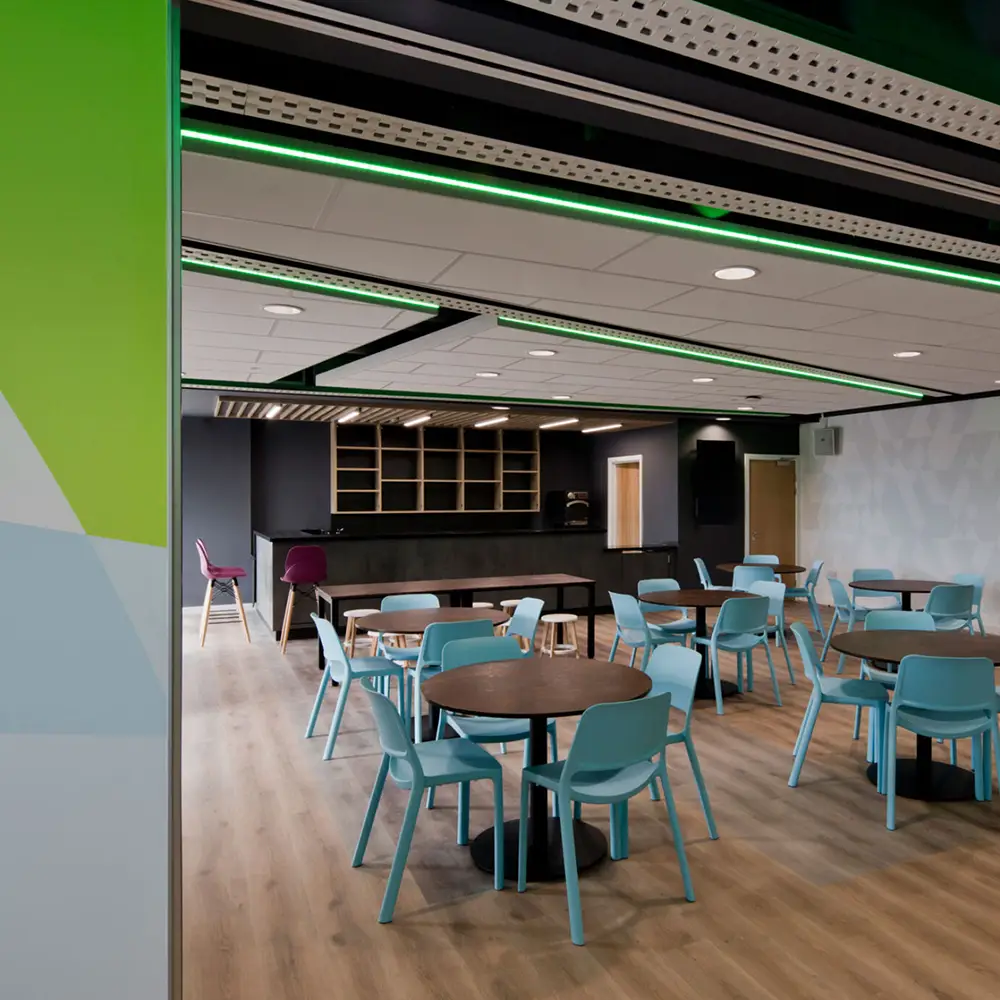 Modern cafeteria with round dark tables and light blue chairs on wood flooring under a ceiling with green LED lights.