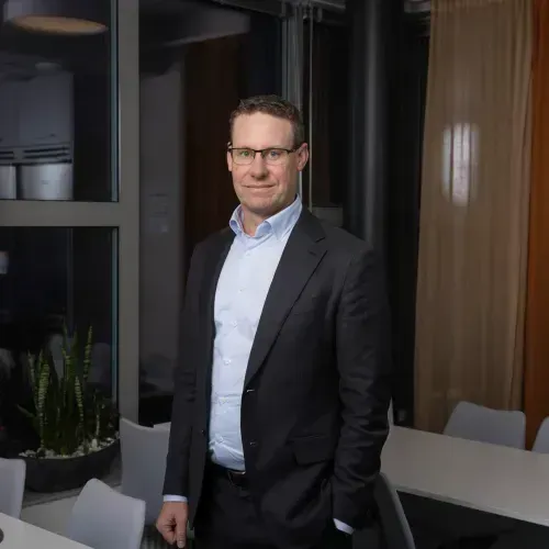 Man wearing glasses and a suit standing in a modern office near a window and table with chairs.
